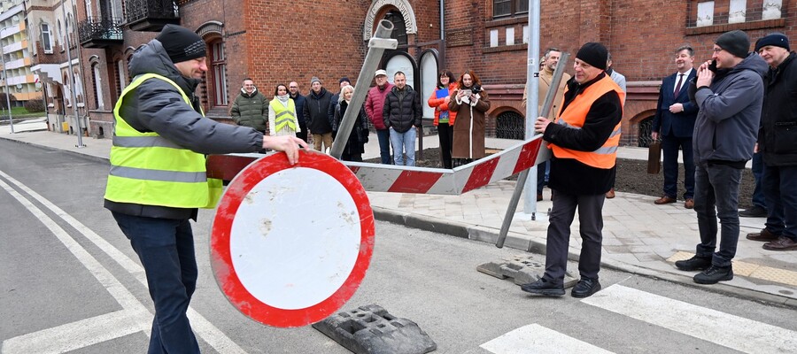 dwóch wykonawców przenosi szlaban, w tle uczestnicy potkania